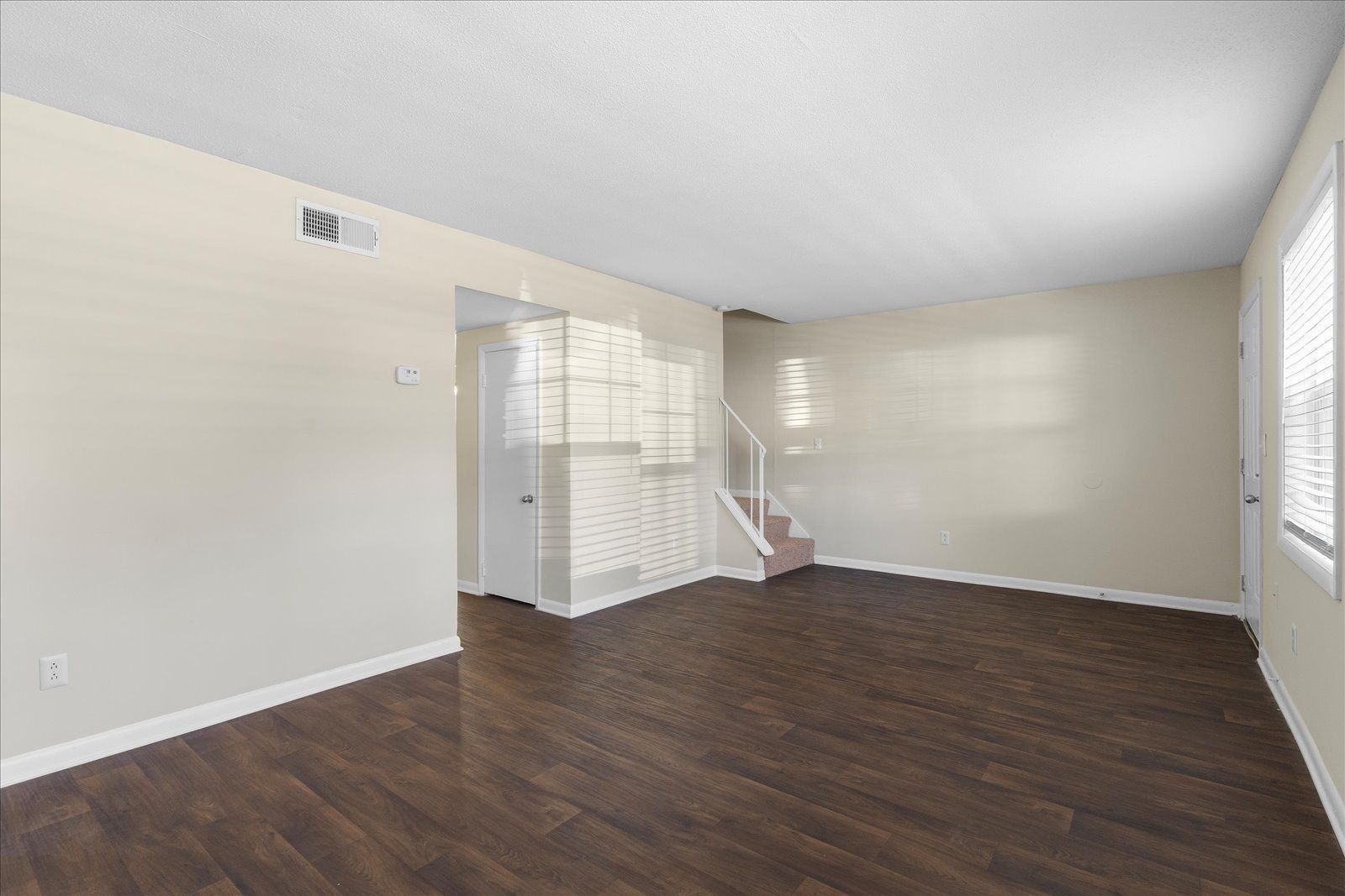 Empty living room with dark wood floors, beige walls, and white blinds on windows.