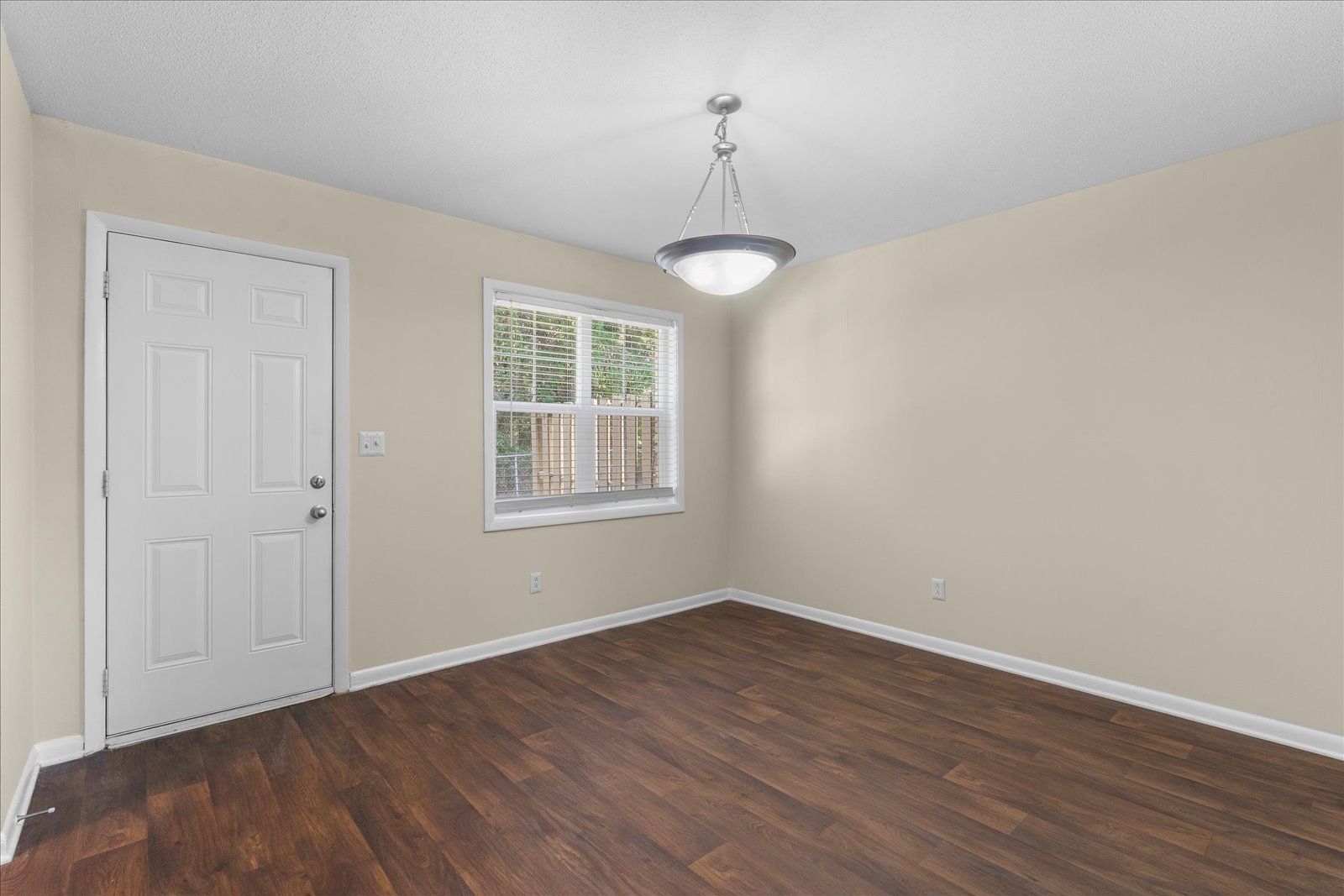 Empty beige room with dark wood floors, white door, window, and ceiling light.