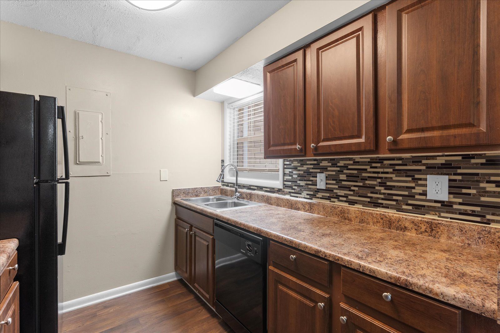 Compact kitchen with dark wood cabinets, speckled countertop, black appliances, and mosaic tile backsplash.