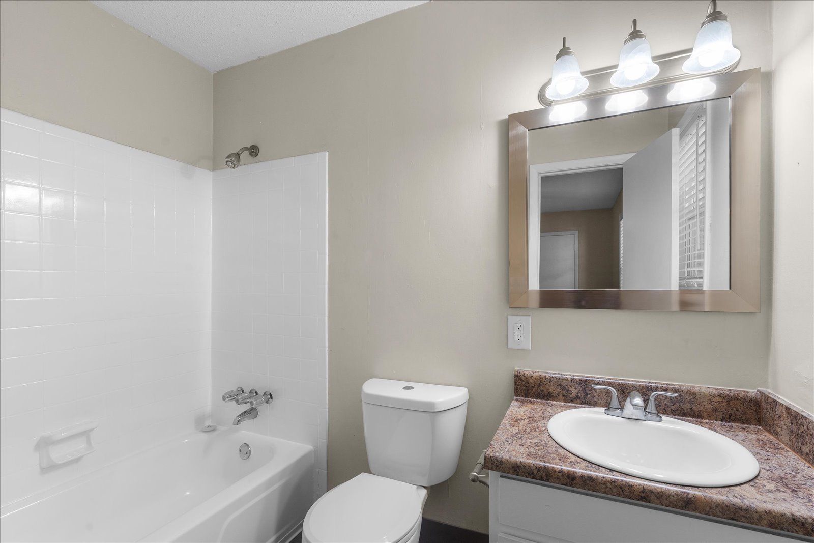 Small bathroom with white tub, toilet, and vanity with granite countertop and mirror lights.