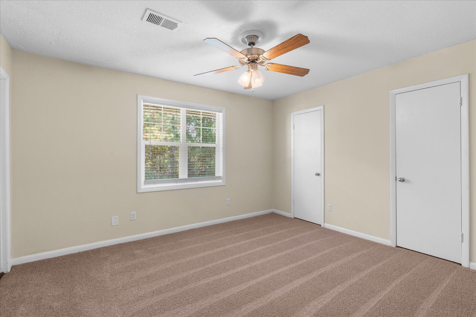Empty beige bedroom with carpet, ceiling fan, window, and two white doors