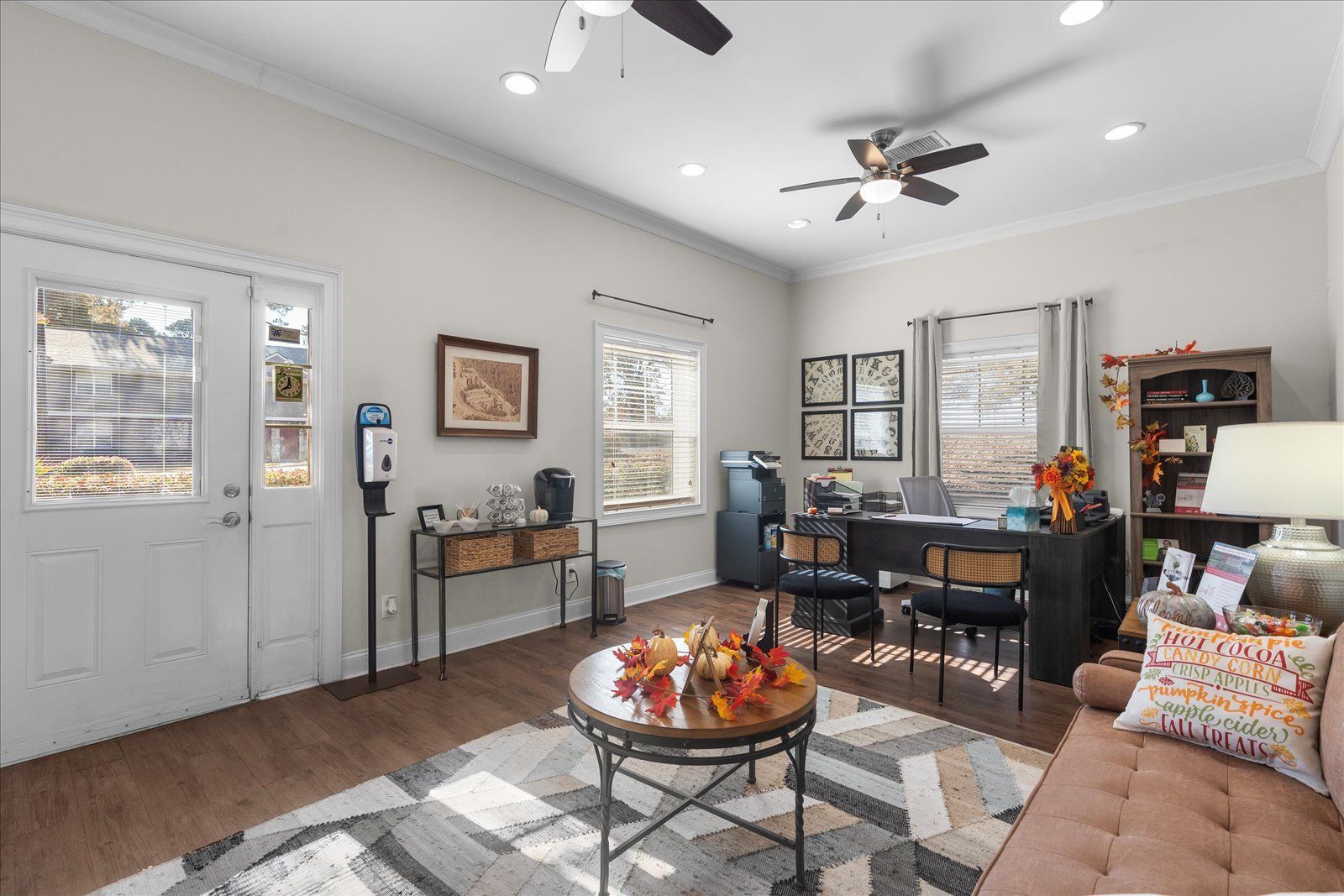 Bright living room with white walls, gray sofa, coffee table, desk, and ceiling fan, lit by windows.