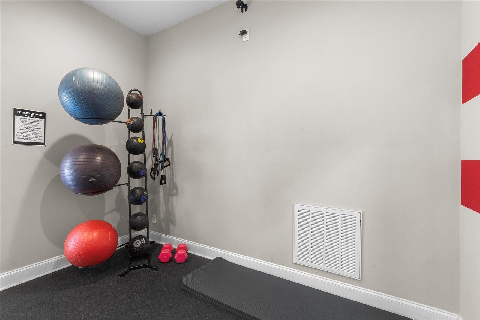Gym corner with exercise balls, medicine-ball rack, pink dumbbells, and a wall vent on gray walls.