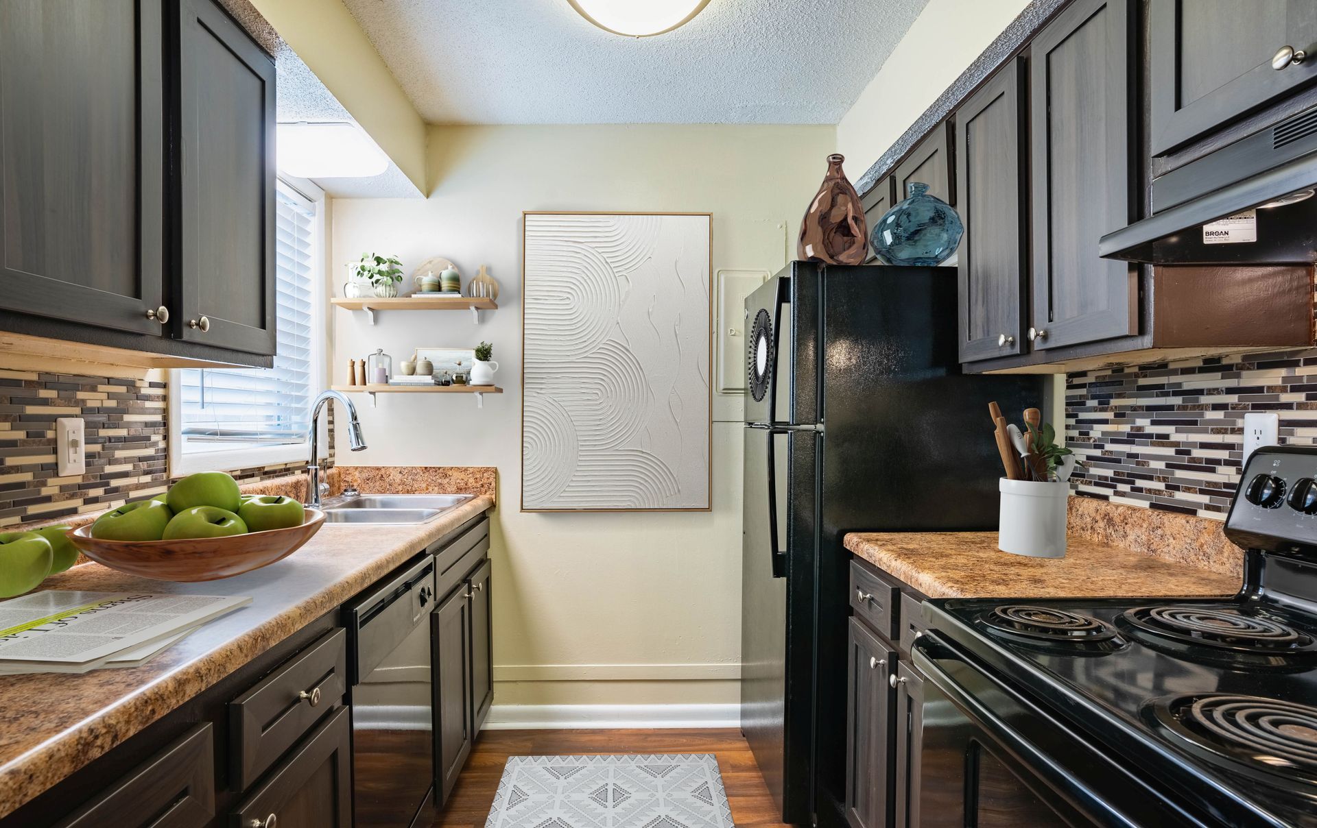 Narrow kitchen with dark cabinets, black appliances, mosaic backsplash, and a white door at the end.