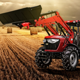 Red tractor with front loader lifting hay bales in a harvested field at sunset