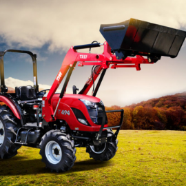 Red tractor with front loader lifting a black bucket in a field with autumn trees in the background