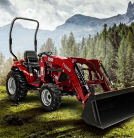 Red tractor with front loader on a grassy mountain path, forest and misty peaks in the background