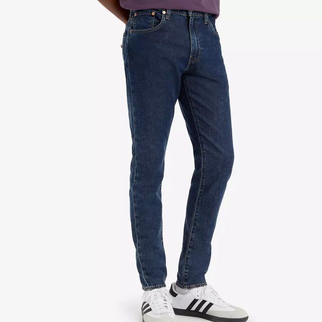 Un uomo che indossa una maglietta viola e jeans blu è in piedi su uno sfondo bianco.