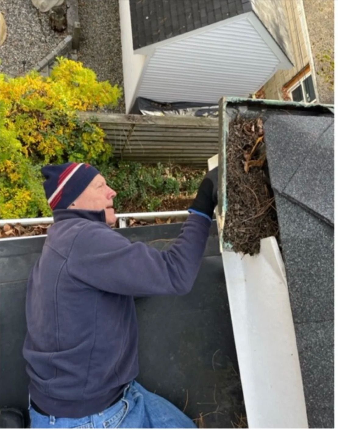Person cleaning a gutter or roof edge beside a house, wearing a beanie and hoodie