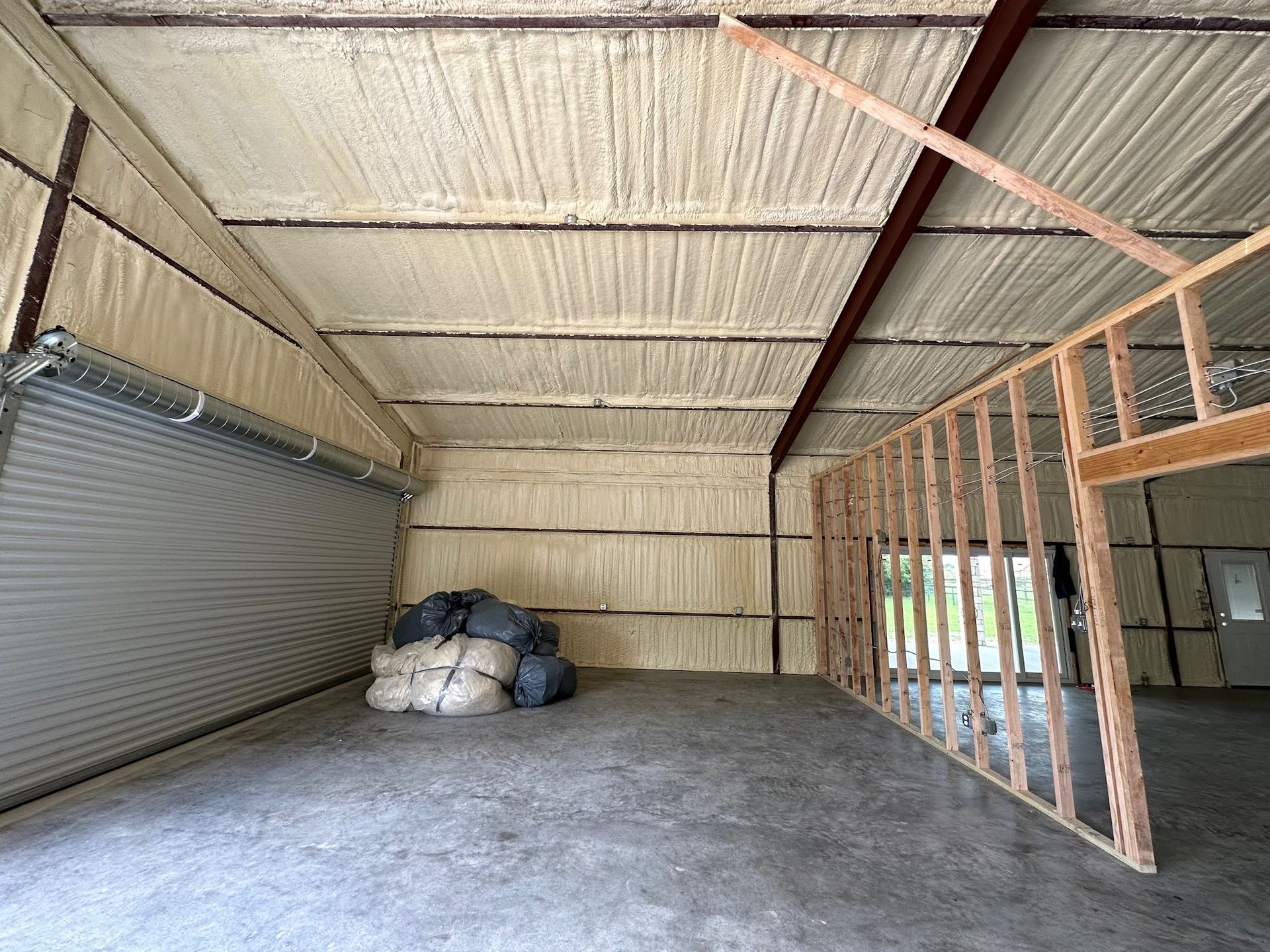 Interior of a metal building with spray foam insulation. A garage door and framed wall are visible.