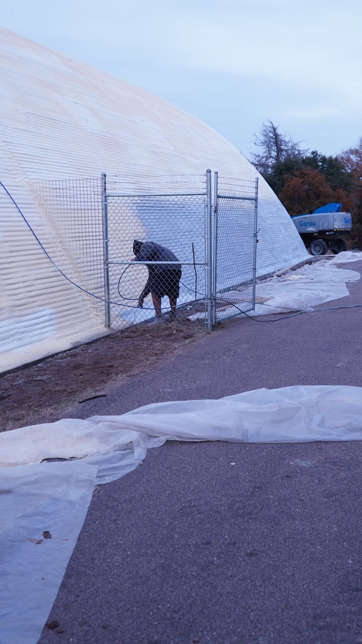 Person shoveling snow near a white dome structure behind a chain-link fence.