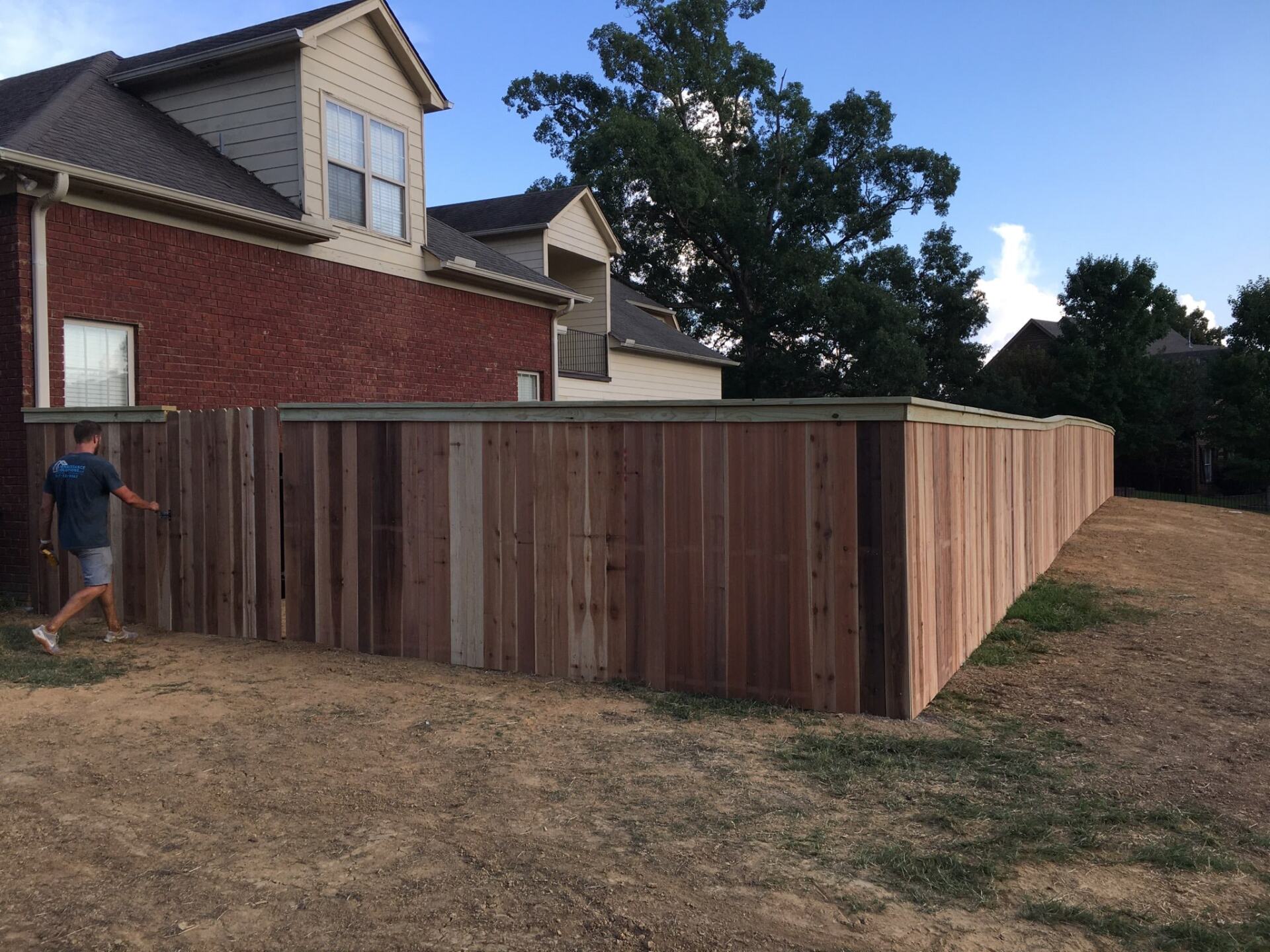 A man is walking behind a wooden fence in front of a brick house.