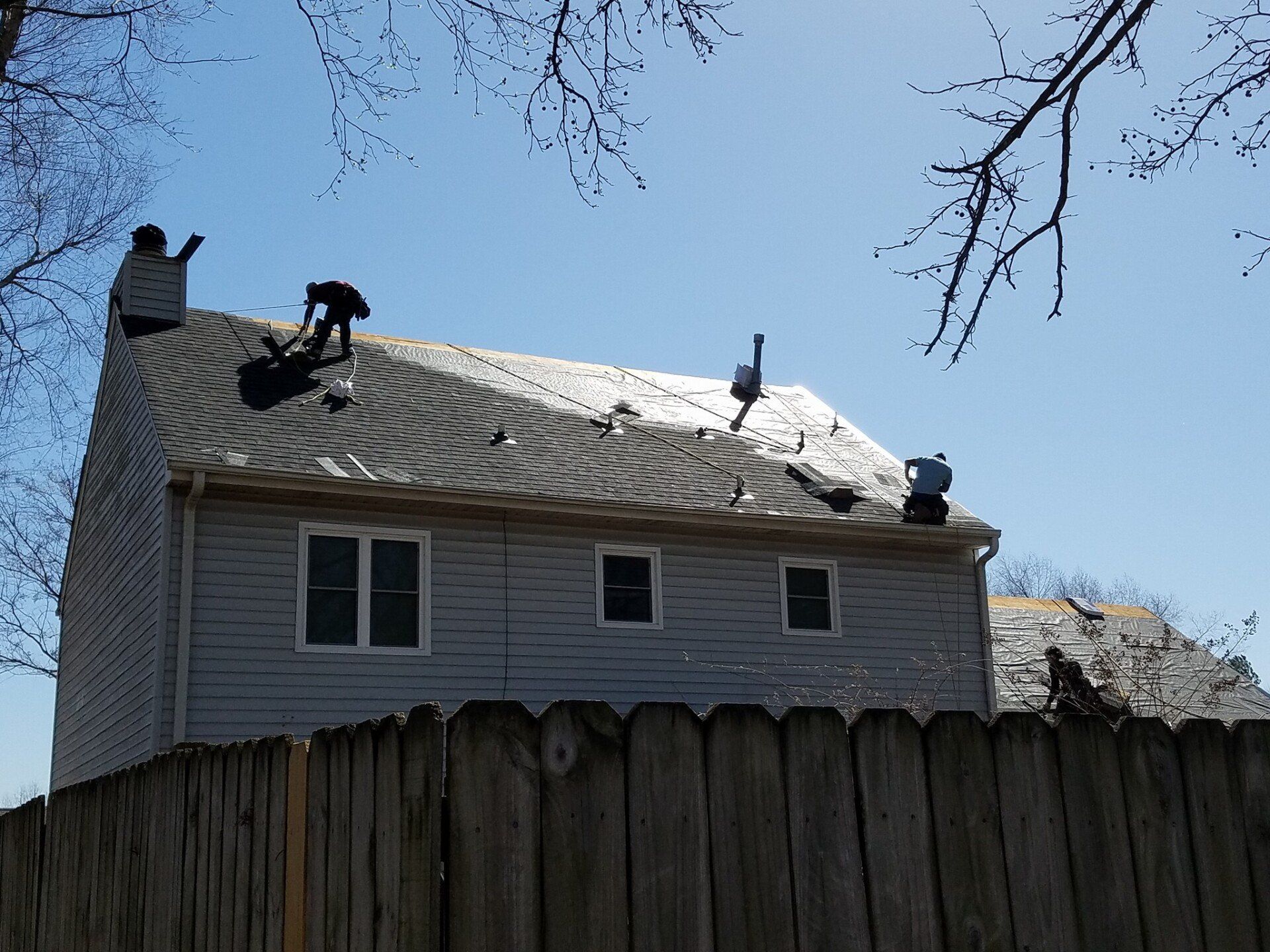 Two men are working on the roof of a house next to a wooden fence.