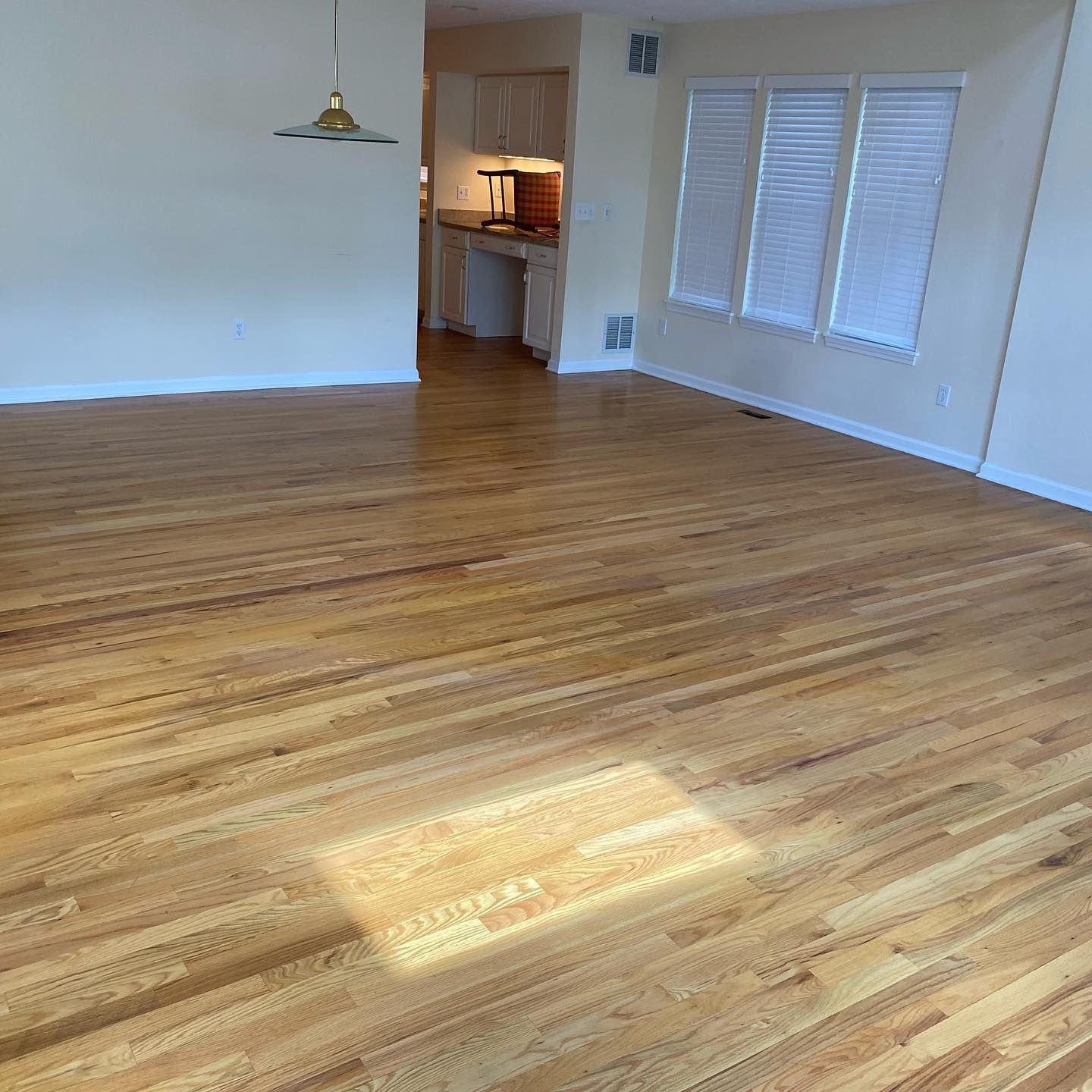 A bright, empty room with polished hardwood floors and three white-blinded windows, looking into a kitchen area.