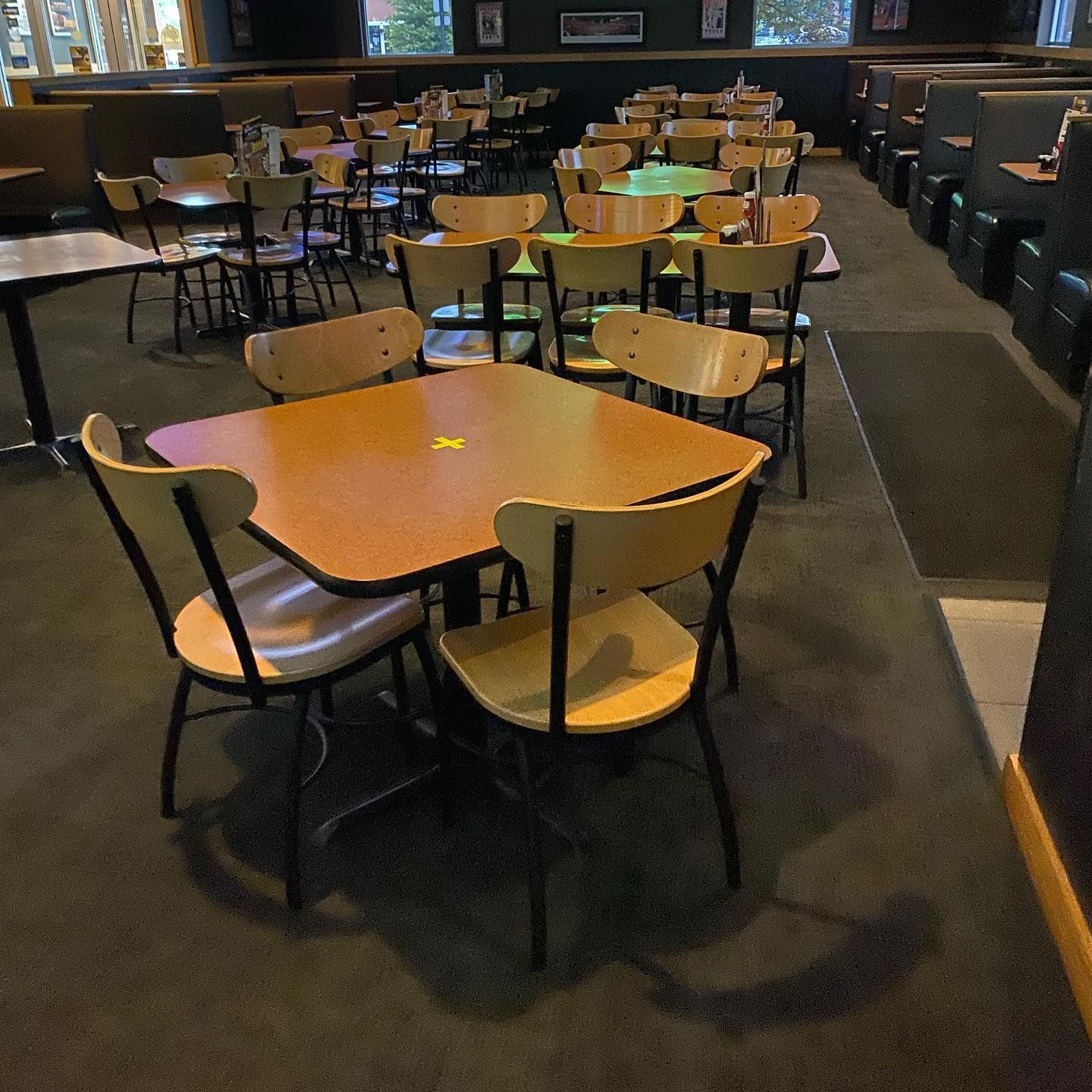 A dimly lit restaurant interior with many empty square tables and wooden chairs arranged in rows on a dark carpet.