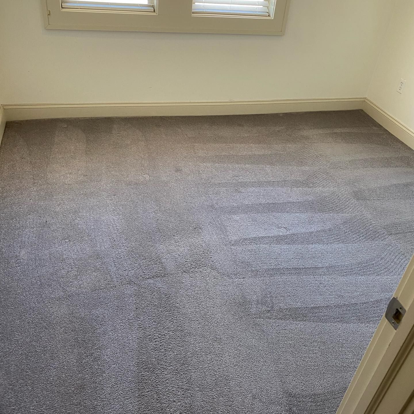 An empty room with gray, textured wall-to-wall carpeting and off-white walls featuring a window with plantation shutters.