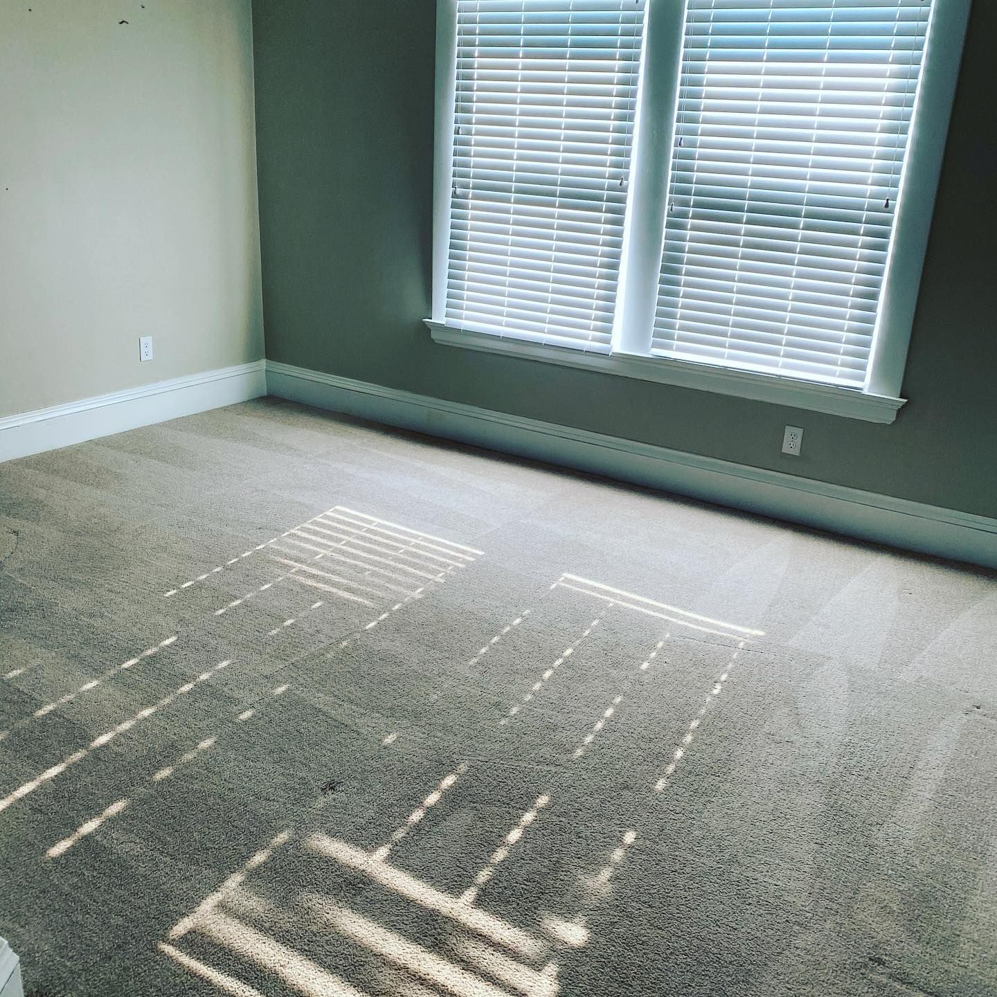 A gray-carpeted room with white baseboards and light streaming through white blinds, casting patterns on the floor.