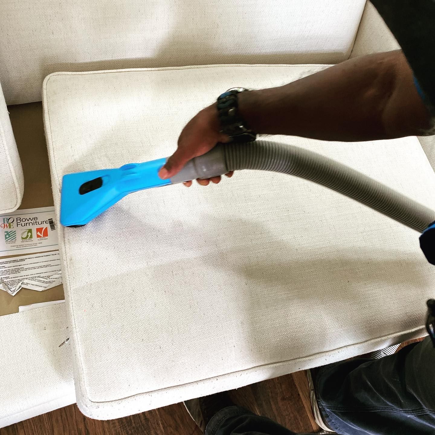 A person uses a blue upholstery cleaning tool to clean a light-colored fabric cushion.