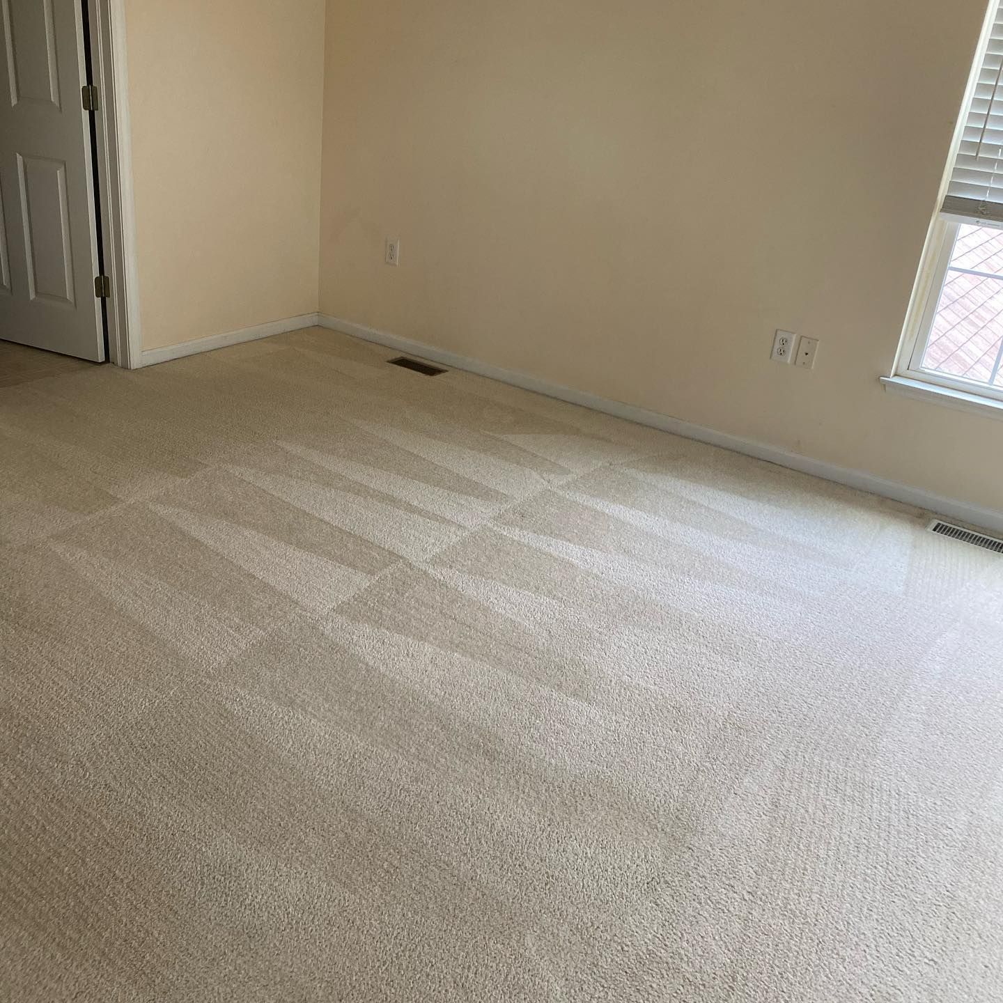 An empty room with beige carpet, cream-colored walls, a white door on the left, and a window with blinds on the right.