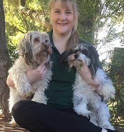 Woman smiling, holding two small dogs. One dog is grey, the other black and white. Outdoors, green background.