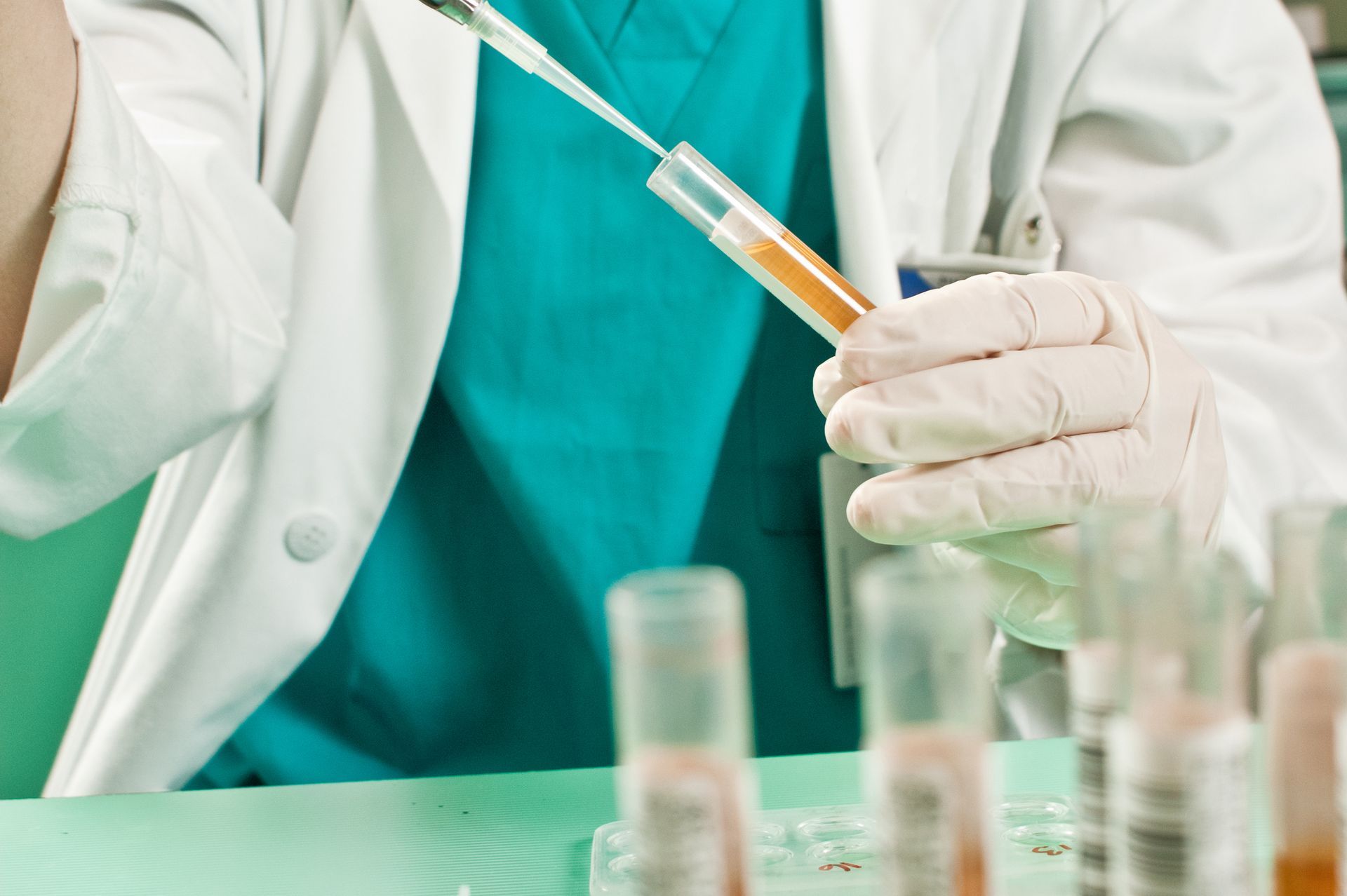 Scientist in lab coat and gloves using a pipette to dispense liquid into a test tube with a yellow liquid.