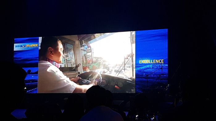 A man is driving a bus in front of a large screen.