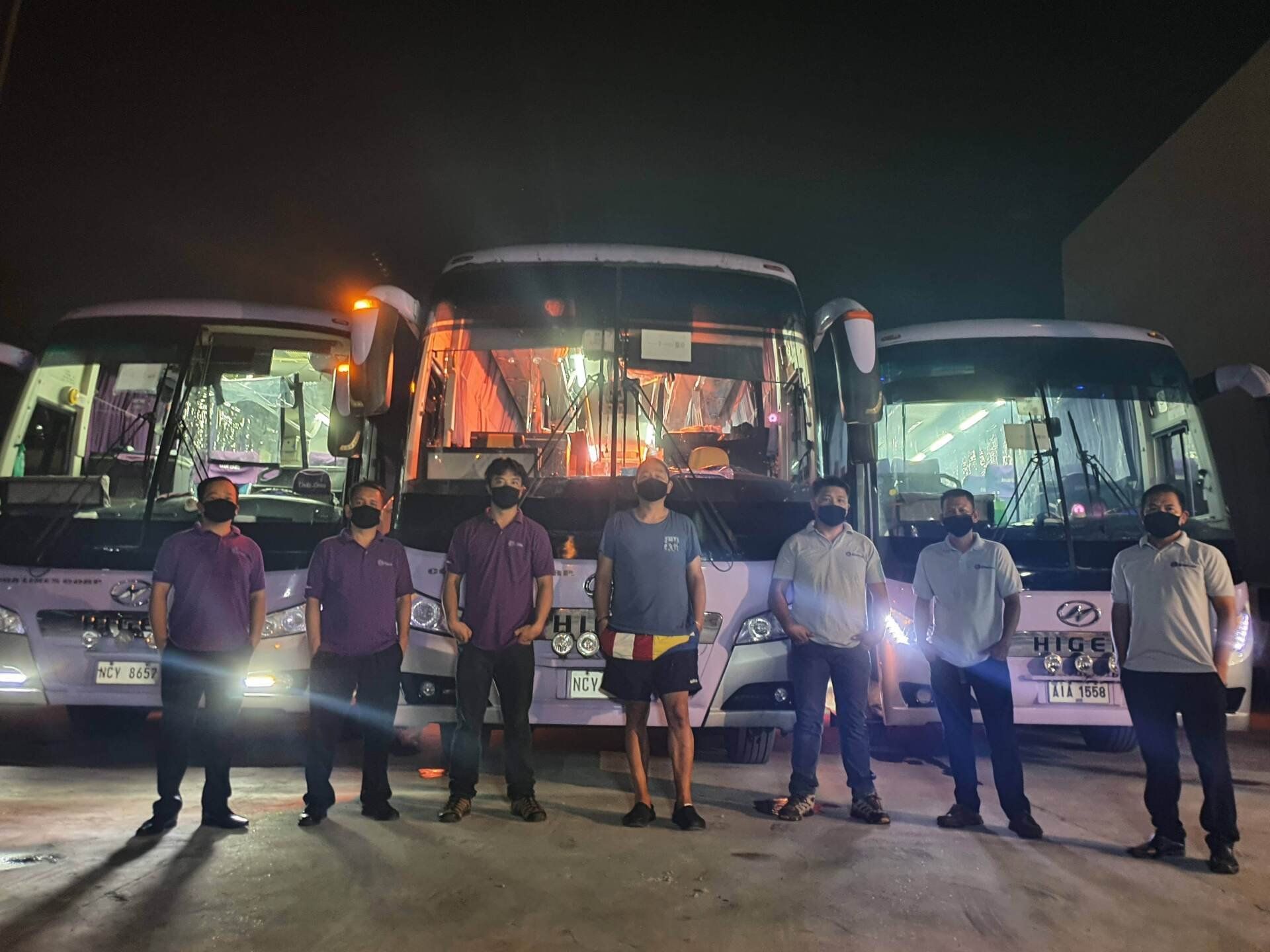 A group of men wearing masks are standing in front of a row of buses.