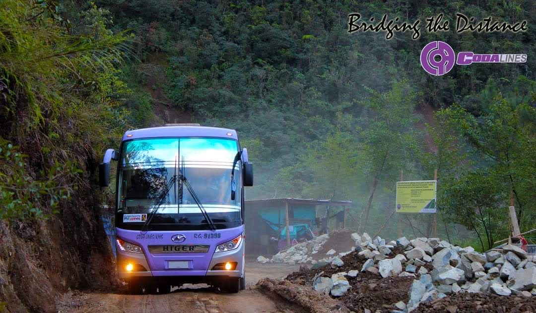 A purple bus is driving down a dirt road.