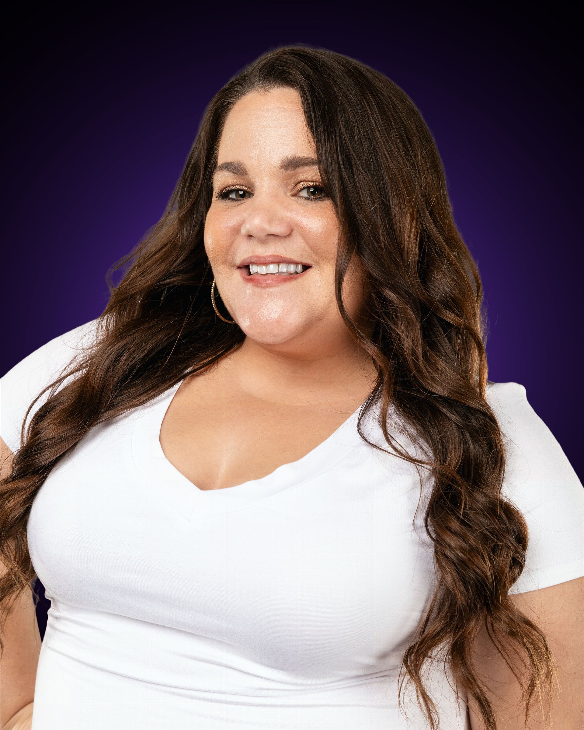 Woman with gray hair wearing a purple shirt, smiling with hands clasped. Set against a gray background.