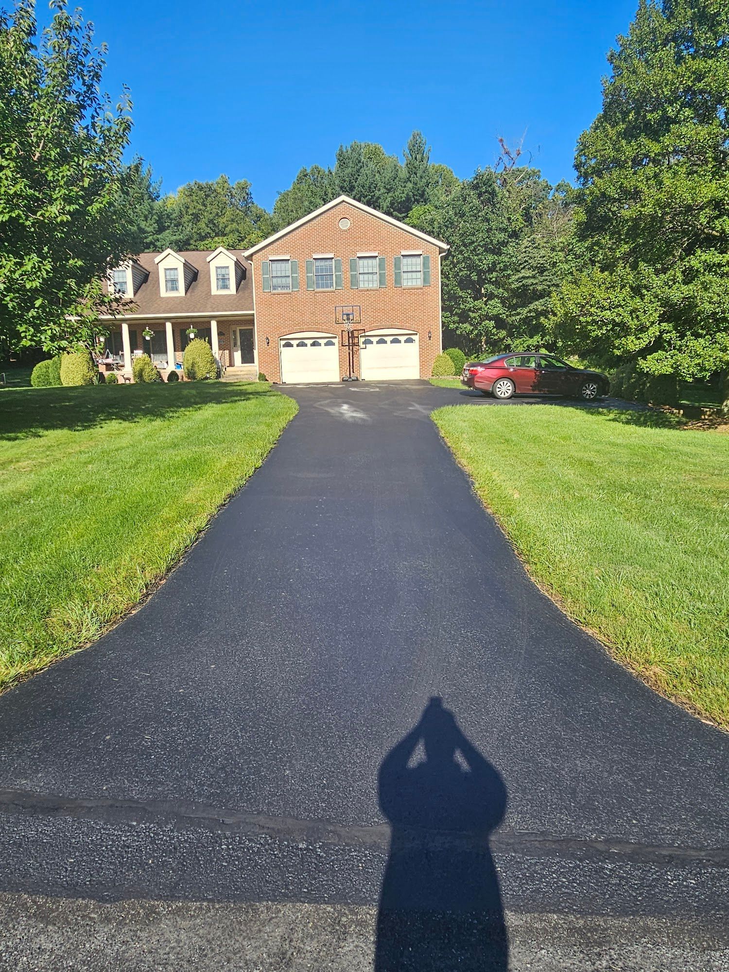 Brick suburban house with double garage, long black driveway, green lawn, and red car under a blue sky