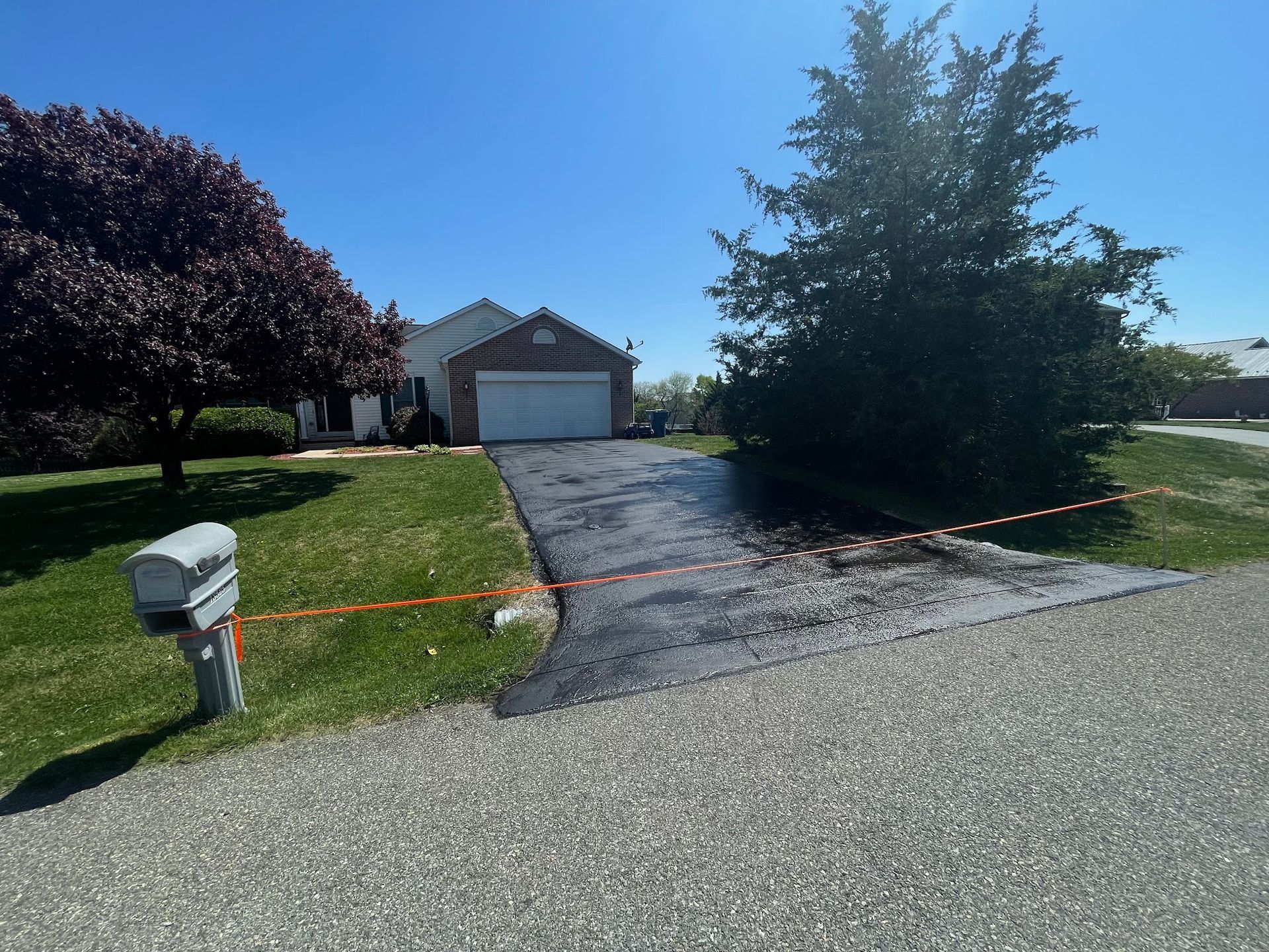Suburban driveway with orange caution tape, a mailbox, trees, and a garage under a clear blue sky