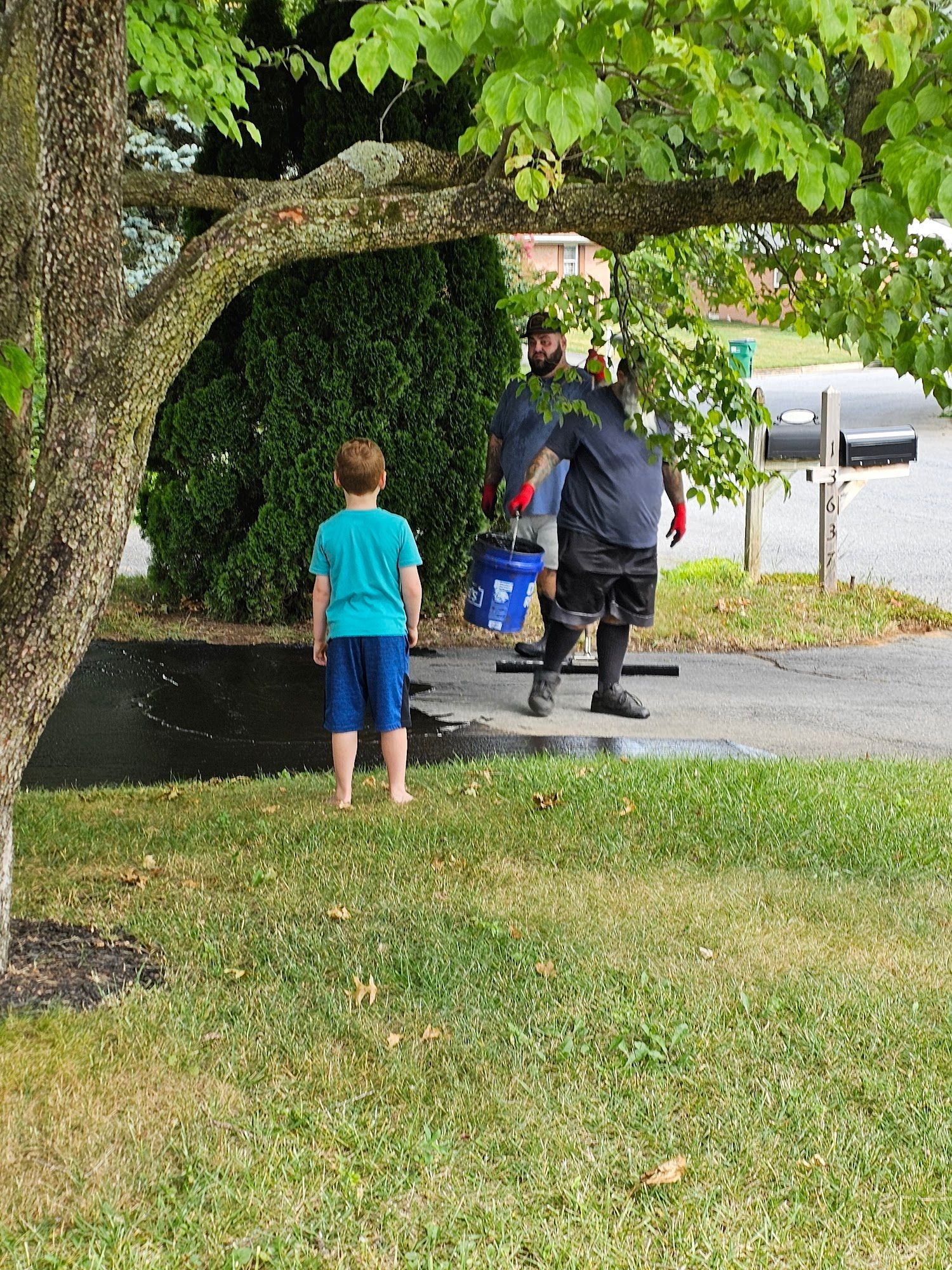 Child in a blue shirt watches a person in black carrying a blue bucket near a driveway under a tree.