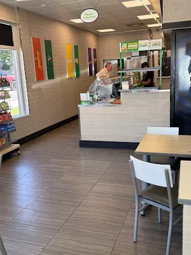 Inside a Subway restaurant: counter, employee preparing food, tables, chairs, and colorful wall decor.