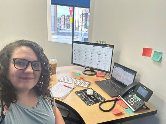 Person smiling at a desk with dual monitors, laptop, phone, and sticky notes in an office.