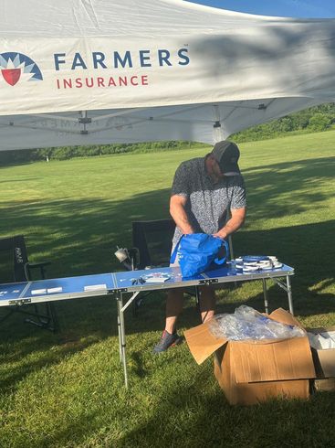 Farmers Insurance booth on a grassy lawn with a person setting up tables and boxes under a white canopy.