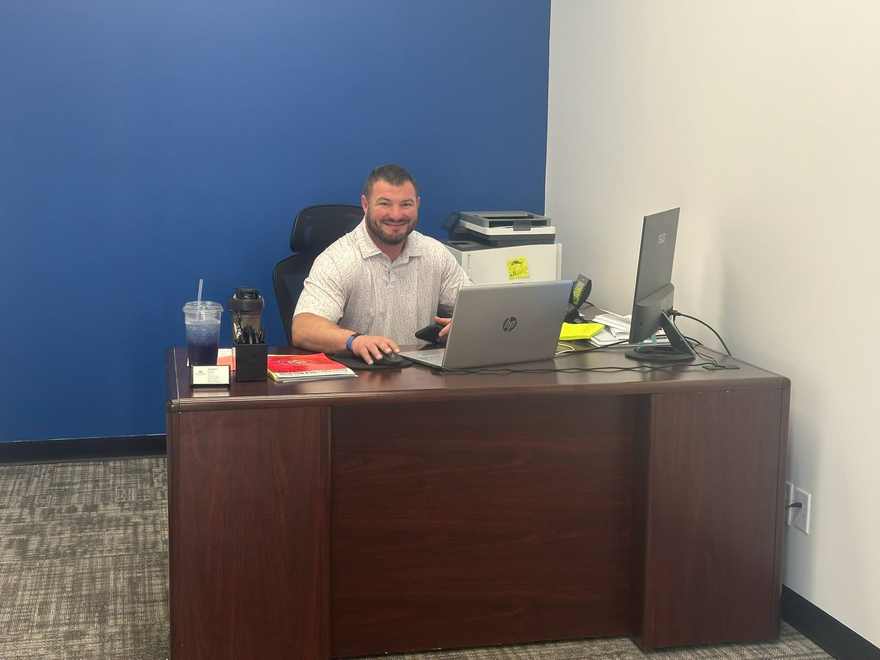 Man sitting at a desk with a laptop in a small office, against a blue wall.