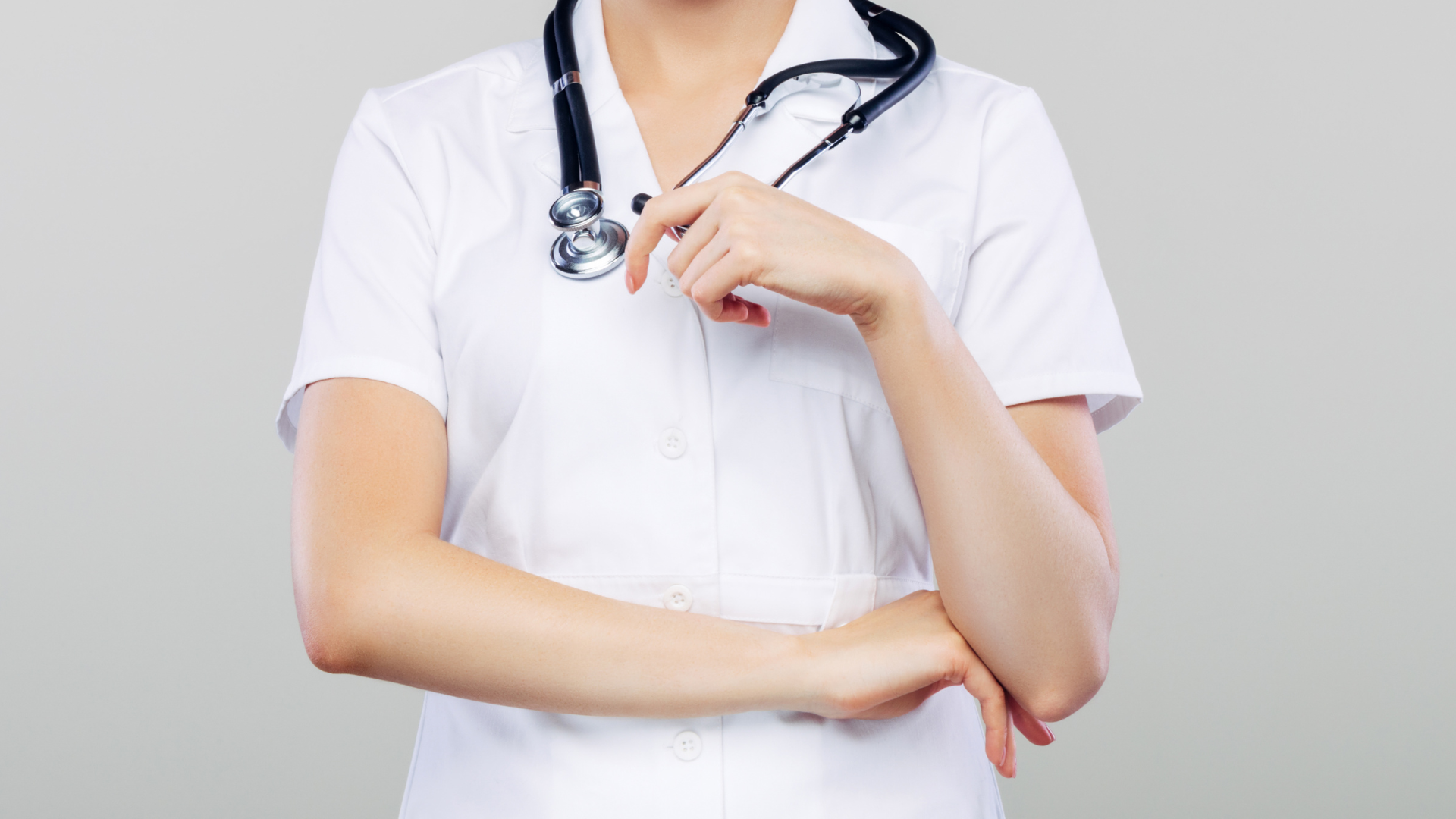 Healthcare worker in white coat with stethoscope, arms crossed against a gray background
