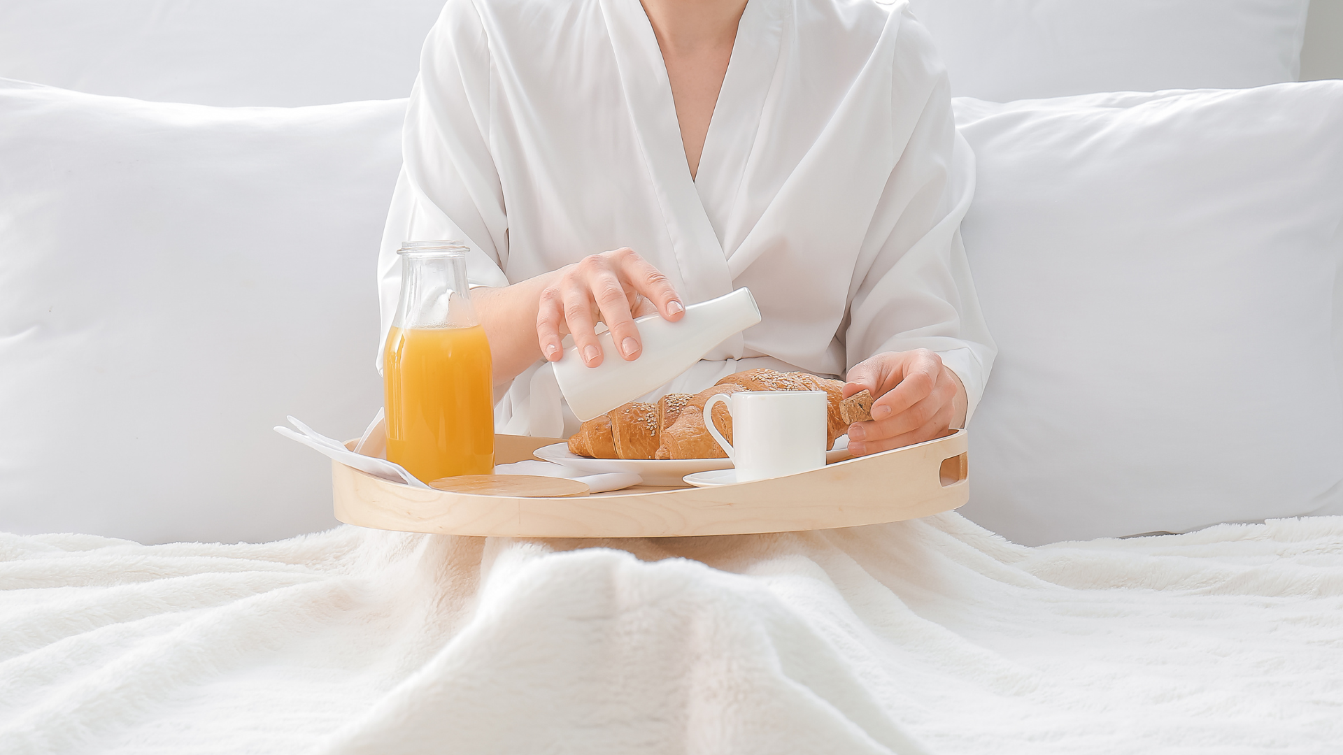 Person in white robe serving breakfast on a bed with orange juice, coffee, and toast on a tray