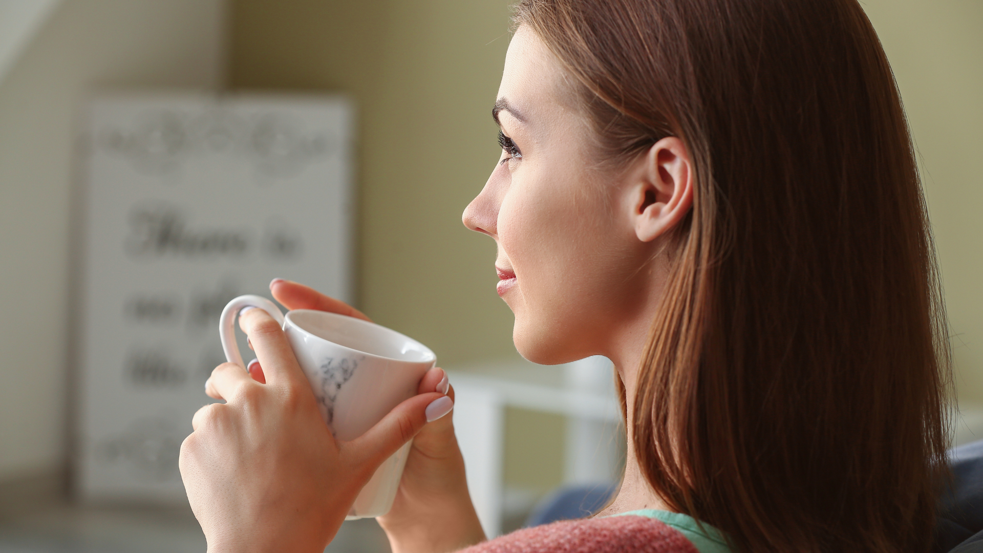 Woman holding a white cup, seen in profile in a softly lit room near a blurred whiteboard.
