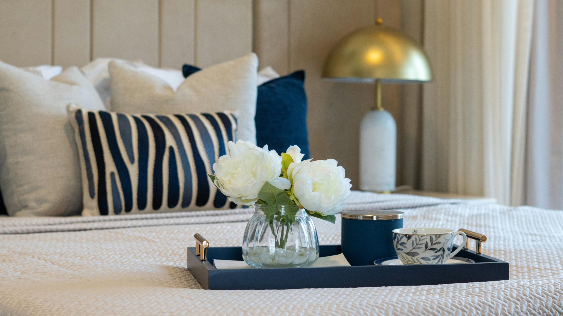 Hotel bed with striped pillows, a tray of white flowers, and a gold lamp on a bedside table