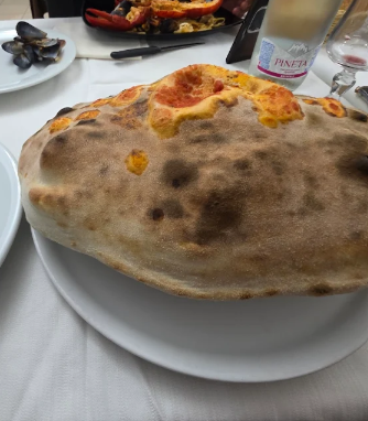 Una grande pizza con la crosta croccante su un piatto bianco su un tavolo, con piatti e un bicchiere sullo sfondo.