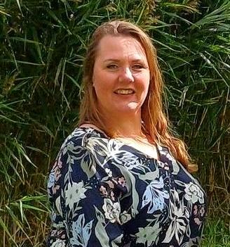 Een vrouw in een bloemetjesblouse staat lachend voor een struik.