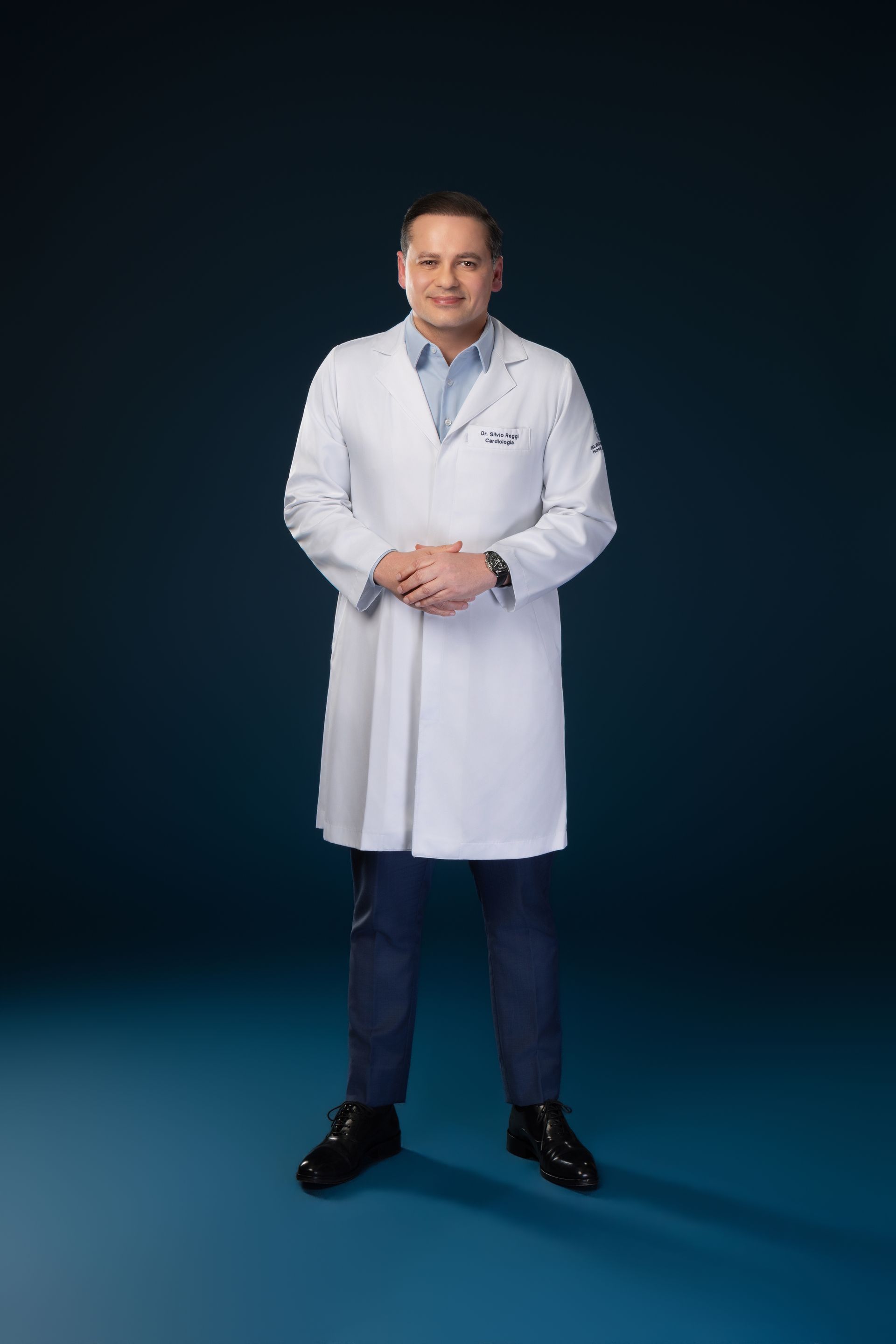 Médico de jaleco branco em pé contra um fundo azul escuro.