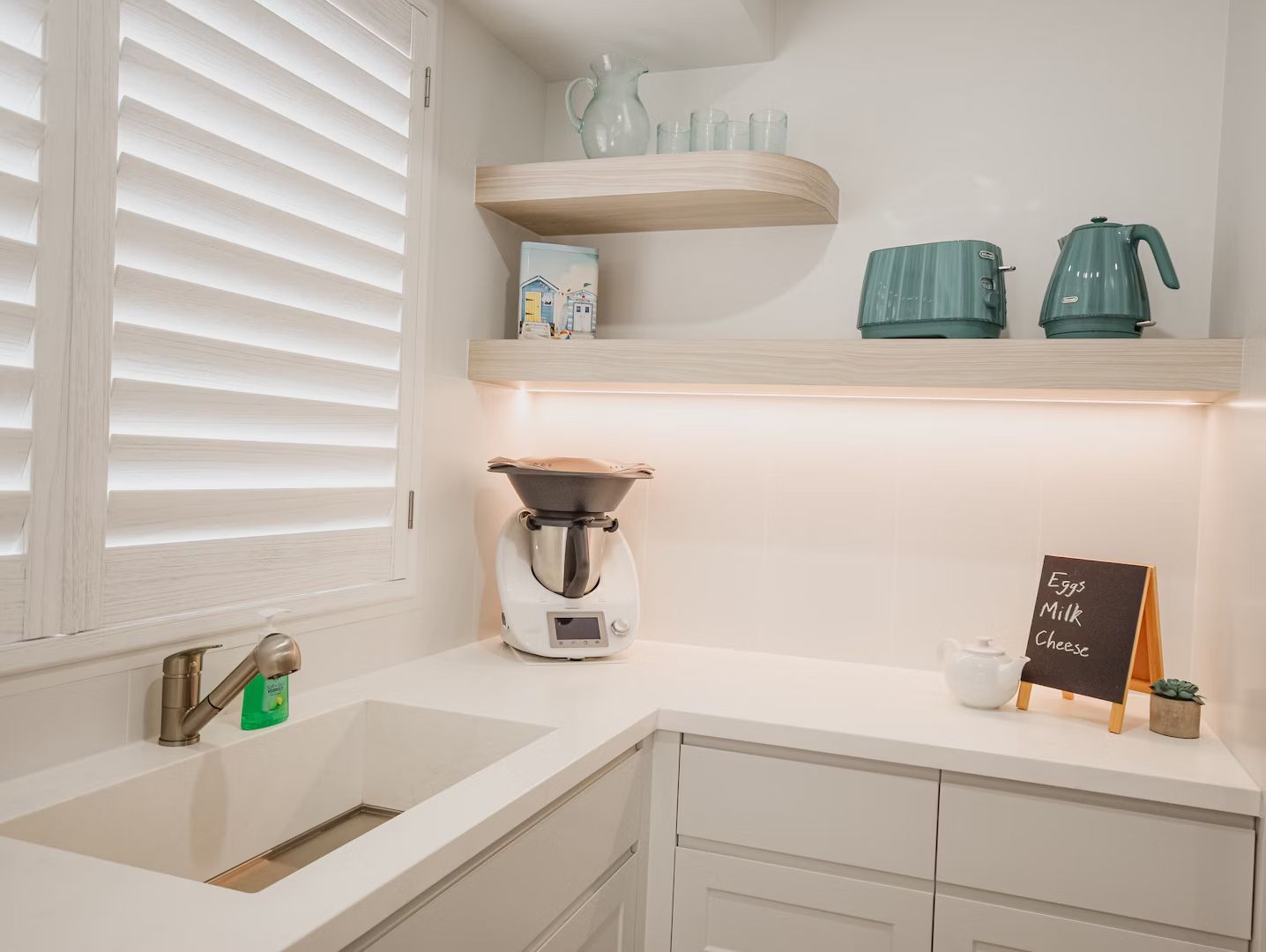 White kitchen with sink, countertop, shelves holding appliances and decor, and shutters.