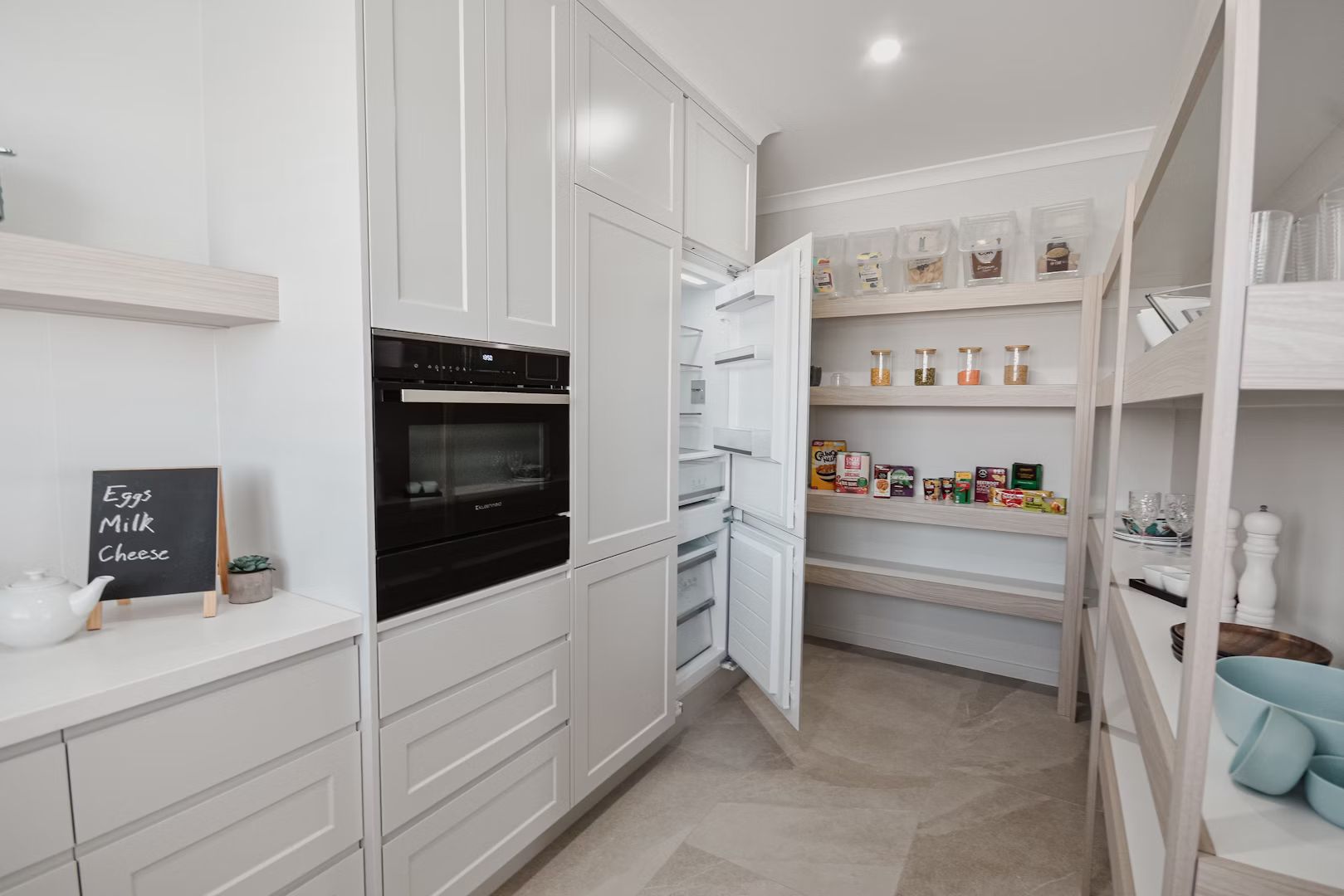 Modern kitchen with open refrigerator, pantry, and built-in oven, all white.