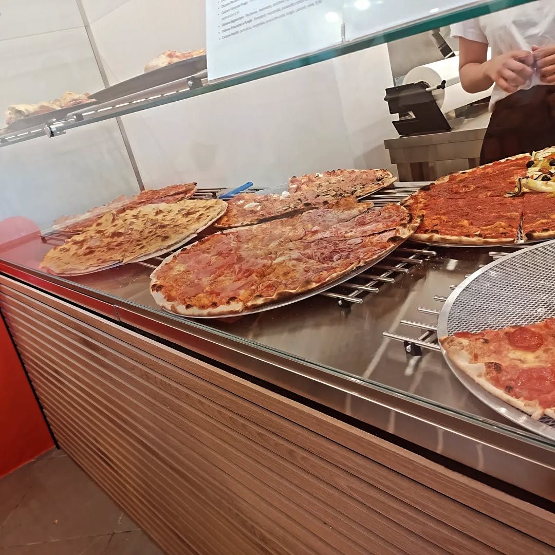 Pizze esposte dietro un bancone di vetro in un ristorante. Sullo sfondo si vede una persona.