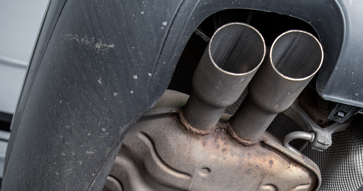 Close-up of a car’s dual exhaust pipes under a rear wheel well, with a rusted muffler. | Leroy's Auto & Truck Care