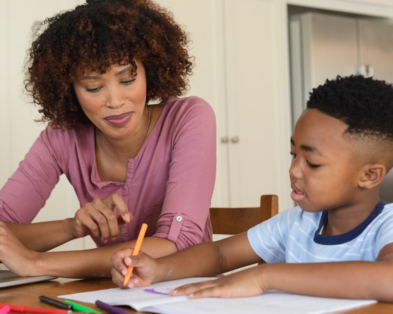 Adult helping child write at a table with pencils and books in a bright room