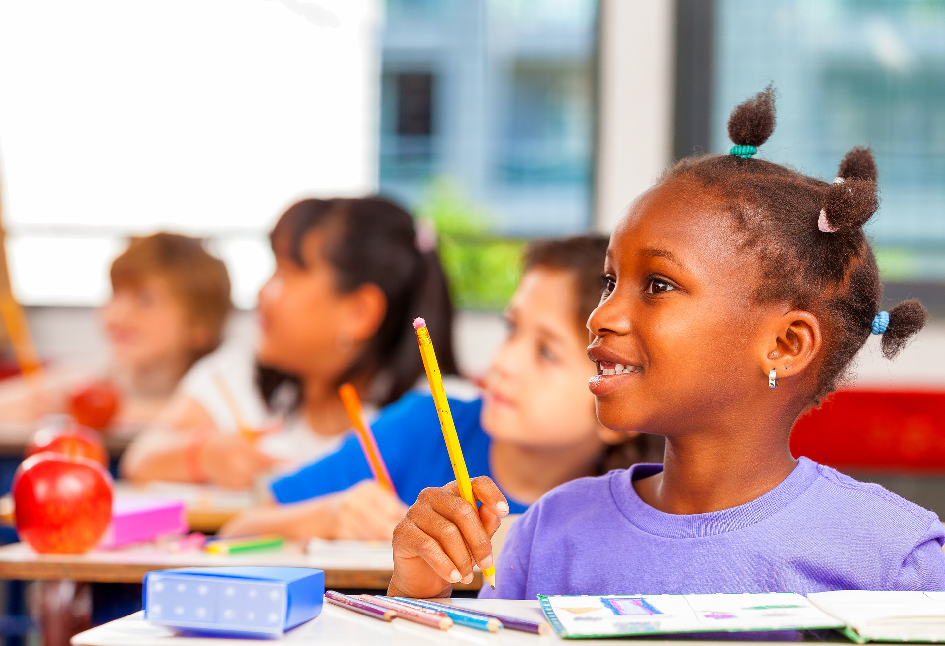 Students sitting at desks in a classroom, one smiling child holding a pencil in the foreground.