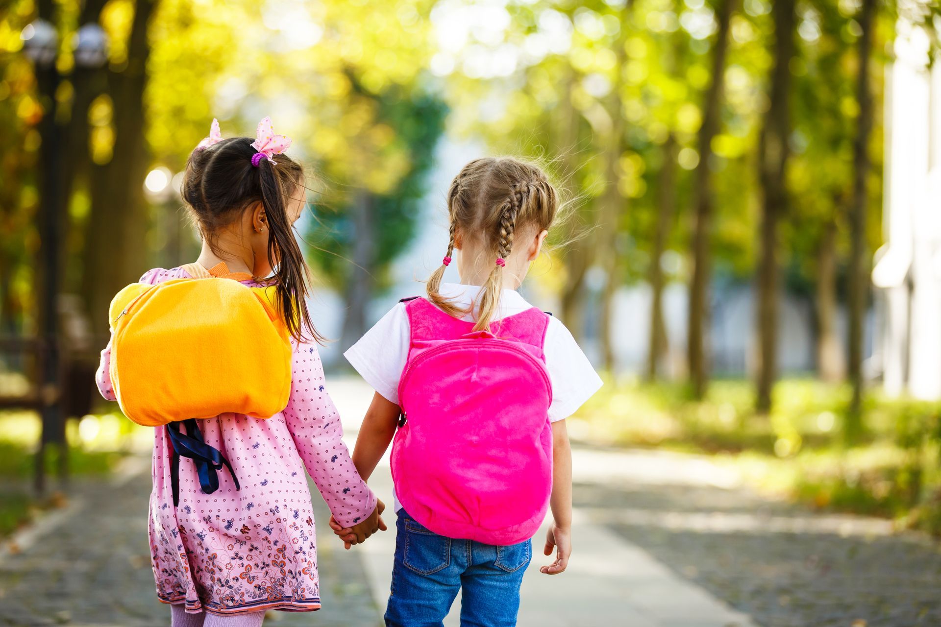 Two children with backpacks holding hands on a sunny park path, seen from behind.